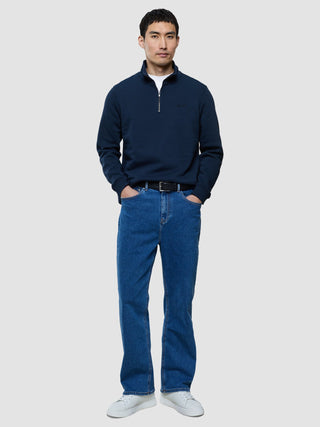 A man wearing a Sweat Half Zip Dark Navy over a white t-shirt, blue jeans with a black belt, and white sneakers.
