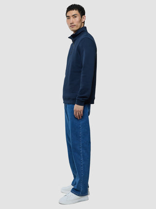 A man stands in profile wearing a Sweat Half Zip Dark Navy, blue jeans, and white sneakers.