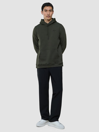 A man stands with his hands behind his back, wearing a Sweat Hoodie Green Night, dark trousers, and white sneakers.