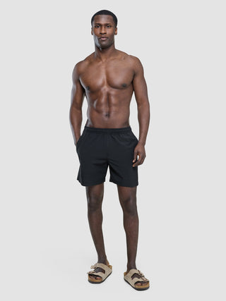 A shirtless man standing and wearing Swim Shorts Black and tan sandals.