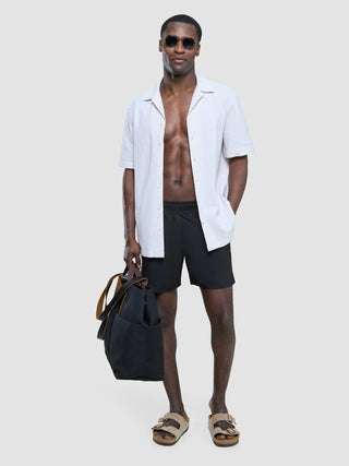 A man wearing sunglasses, an unbuttoned white short-sleeved shirt, Swim Shorts Black, and sandals, holding a dark tote bag.