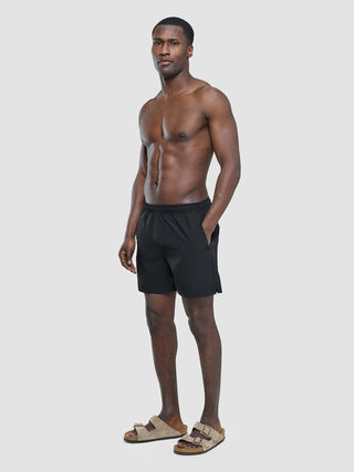 A shirtless man standing and wearing Swim Shorts Black and tan sandals.