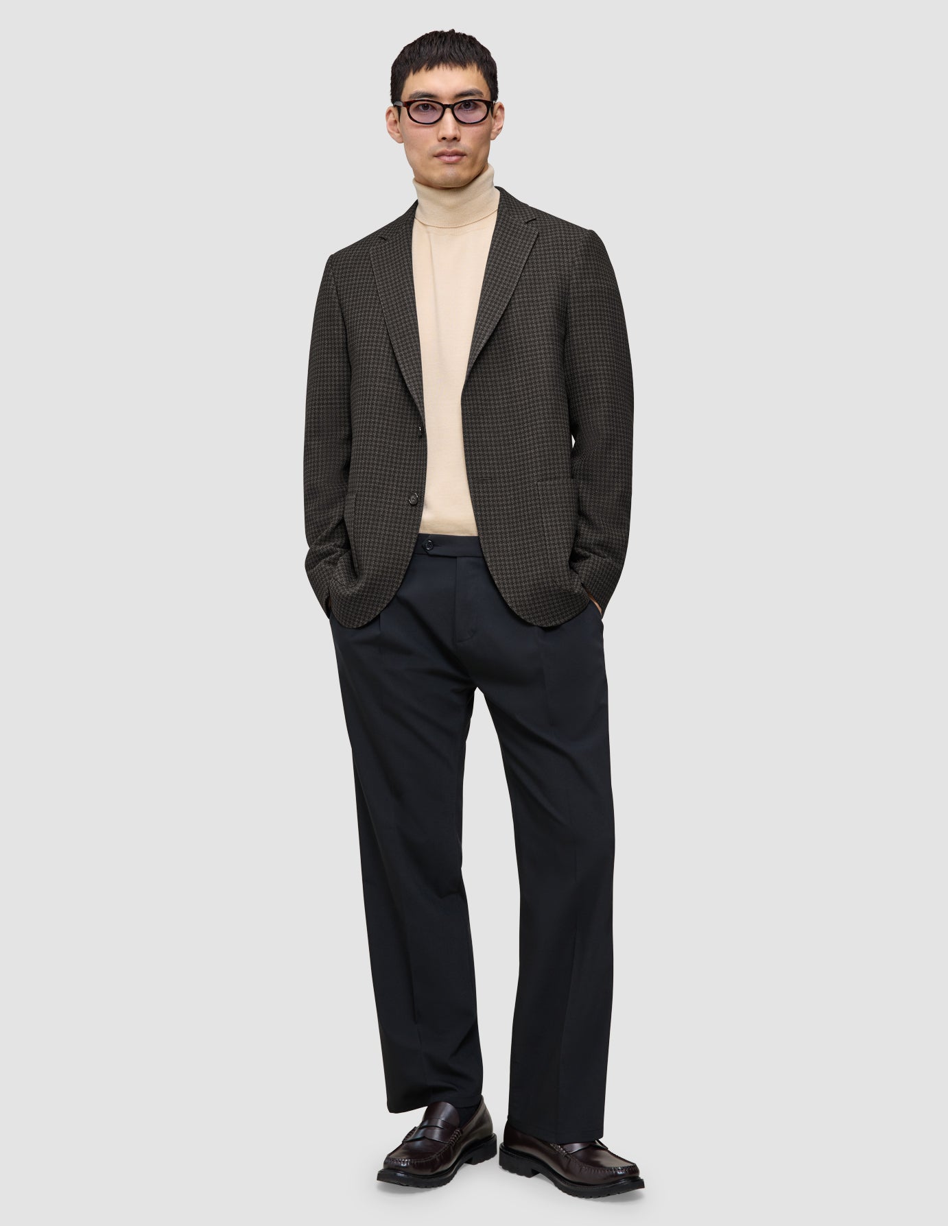 A man wearing a Timeless Wool Blazer Slim Dark Brown Check over a beige turtleneck sweater, dark trousers, and loafers.