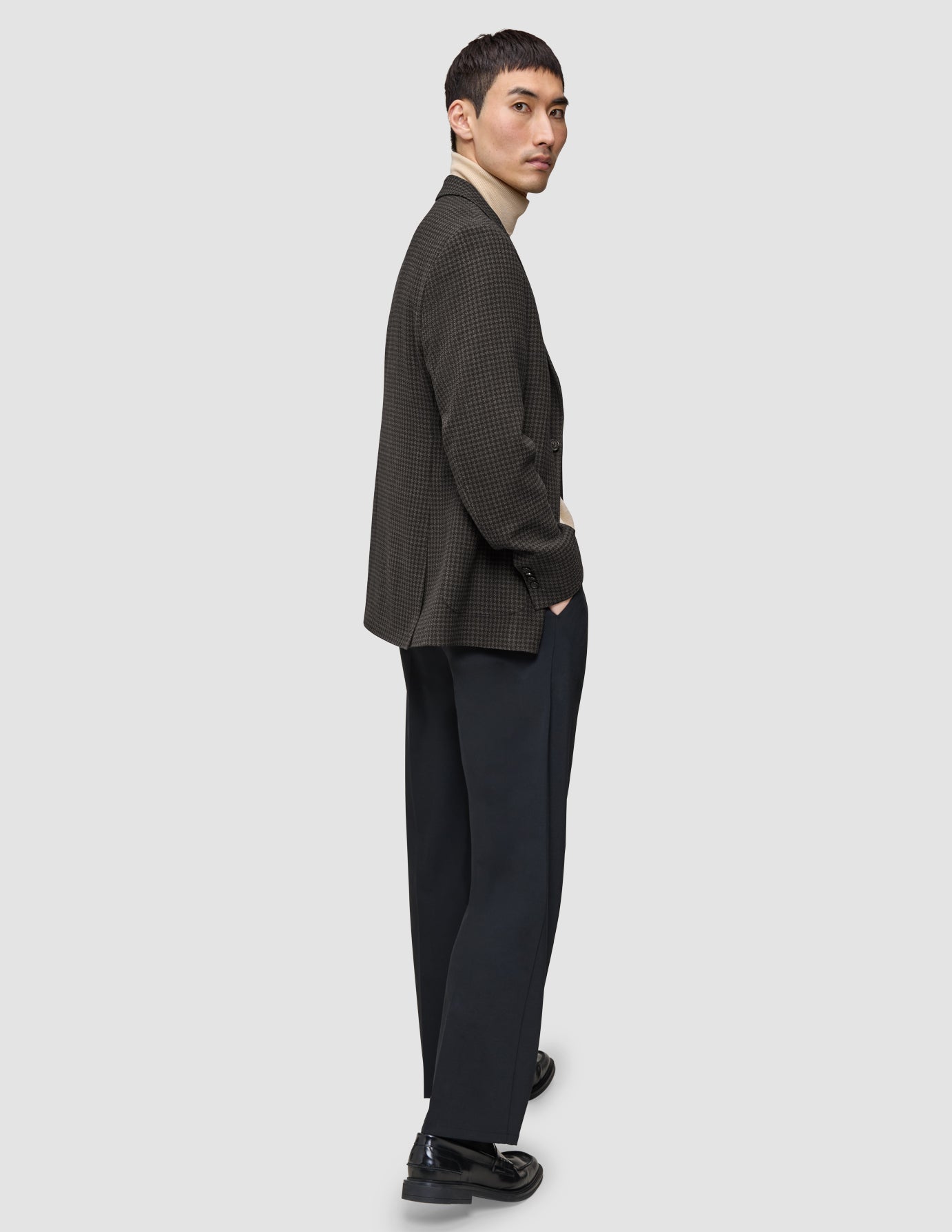 A man wearing a Timeless Wool Blazer Slim Dark Brown Check over a beige turtleneck sweater, paired with dark trousers and black loafers.