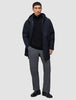 A man wearing a black Utility Jacket Black over a black zip-up sweater, grey trousers, and black shoes.
