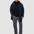 A man wearing a black Utility Jacket Black over a black zip-up sweater, grey trousers, and black shoes.