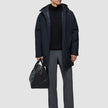 A man wears a black Utility Jacket Black hooded waterproof utility jacket, a black quarter-zip sweater, and grey trousers while holding a black leather duffel bag.