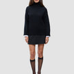 A woman wears a Women Chunky Soft Wool Knit Turtleneck Black over a white collared shirt, paired with a black mini skirt, sheer black stockings, and black pointed-toe heels.