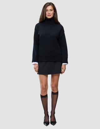 A woman wears a Women Chunky Soft Wool Knit Turtleneck Black over a white collared shirt, paired with a black mini skirt, sheer black stockings, and black pointed-toe heels.