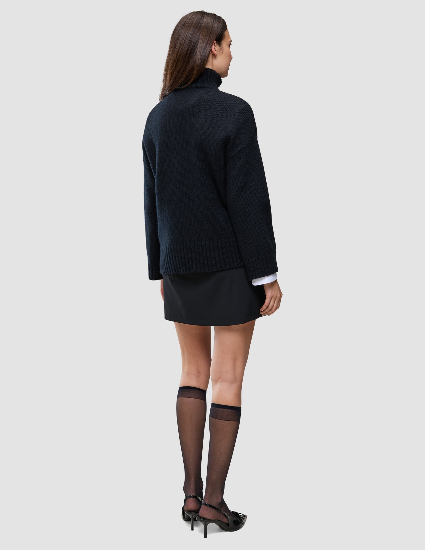 A person seen from behind wearing a Women Chunky Soft Wool Knit Turtleneck Black sweater, a black mini skirt, and sheer black knee-high stockings with black heels.