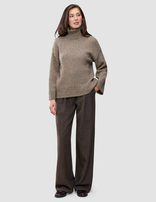 A person wearing a Women Chunky Soft Wool Knit Turtleneck Taupe Melange sweater and dark brown pinstriped trousers.