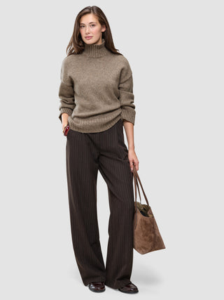 A woman wearing a Women Chunky Soft Wool Knit Turtleneck Taupe Melange sweater and dark pinstriped wide-leg trousers, holding a brown suede tote bag.