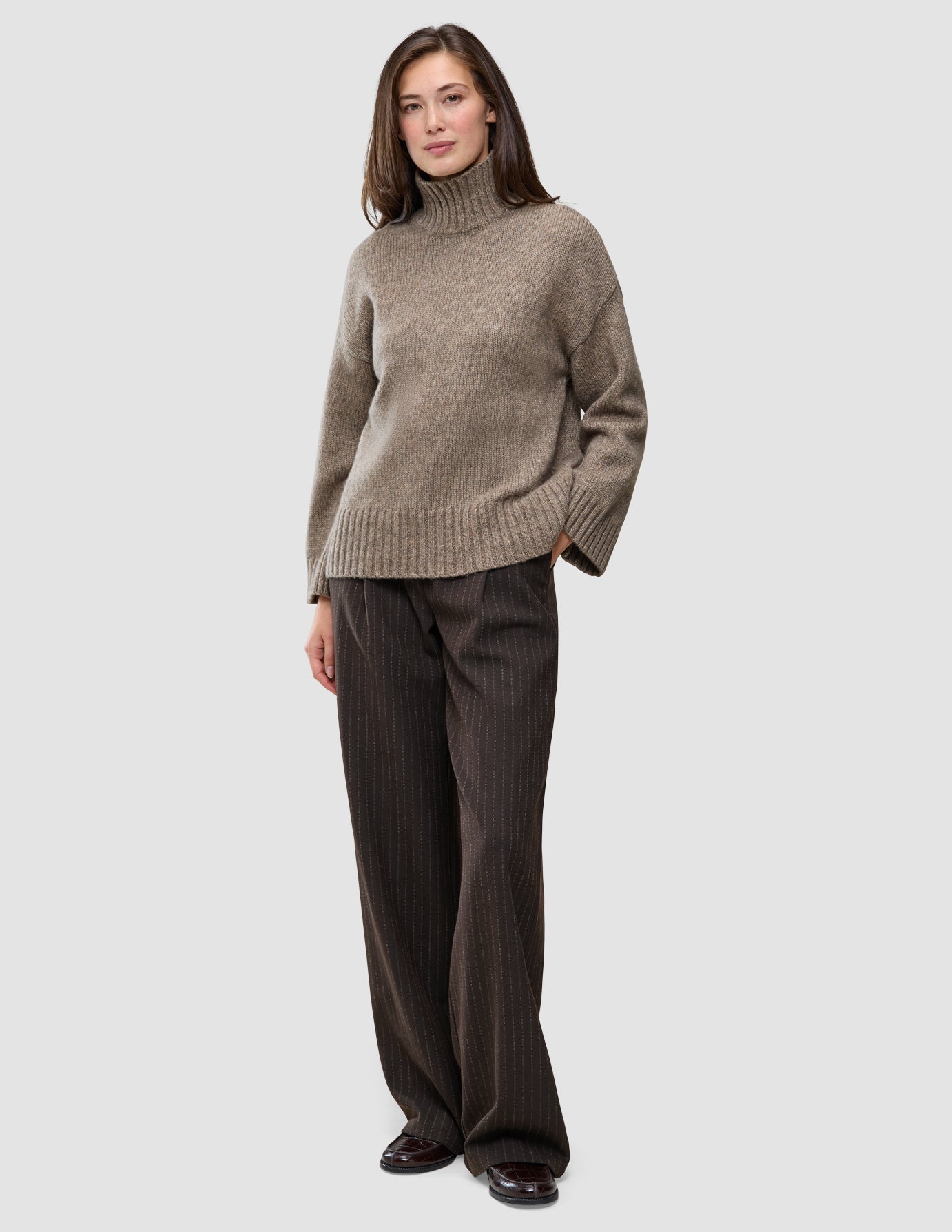 A woman wearing a Women Chunky Soft Wool Knit Turtleneck Taupe Melange in a Scandinavian design with dark brown pinstriped trousers.