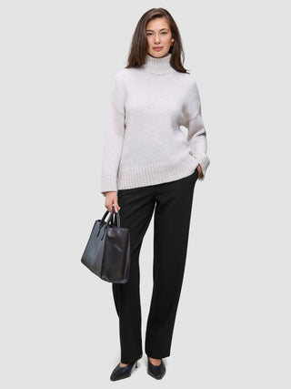 A woman wearing a beige turtleneck sweater and Women GO2 Pants Straight Black, holding a black handbag.