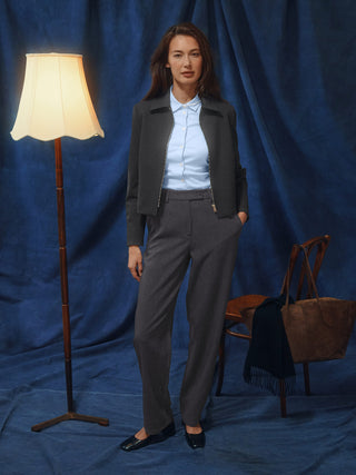 A woman stands wearing a light blue button-down shirt, a dark gray jacket, and Women GO2 Pants Straight Dark Grey Melange, with a floor lamp and a chair behind her.