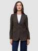 A woman wears a brown Women Serene Blazer Regular Walnut Herringbone with a faint pinstripe pattern, a white t-shirt, and blue jeans, reflecting a clean Scandinavian design.