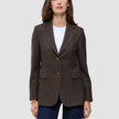 A woman wears a brown Women Serene Blazer Regular Walnut Herringbone with a faint pinstripe pattern, a white t-shirt, and blue jeans, reflecting a clean Scandinavian design.