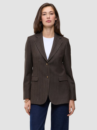A woman wears a brown Women Serene Blazer Regular Walnut Herringbone with a faint pinstripe pattern, a white t-shirt, and blue jeans, reflecting a clean Scandinavian design.