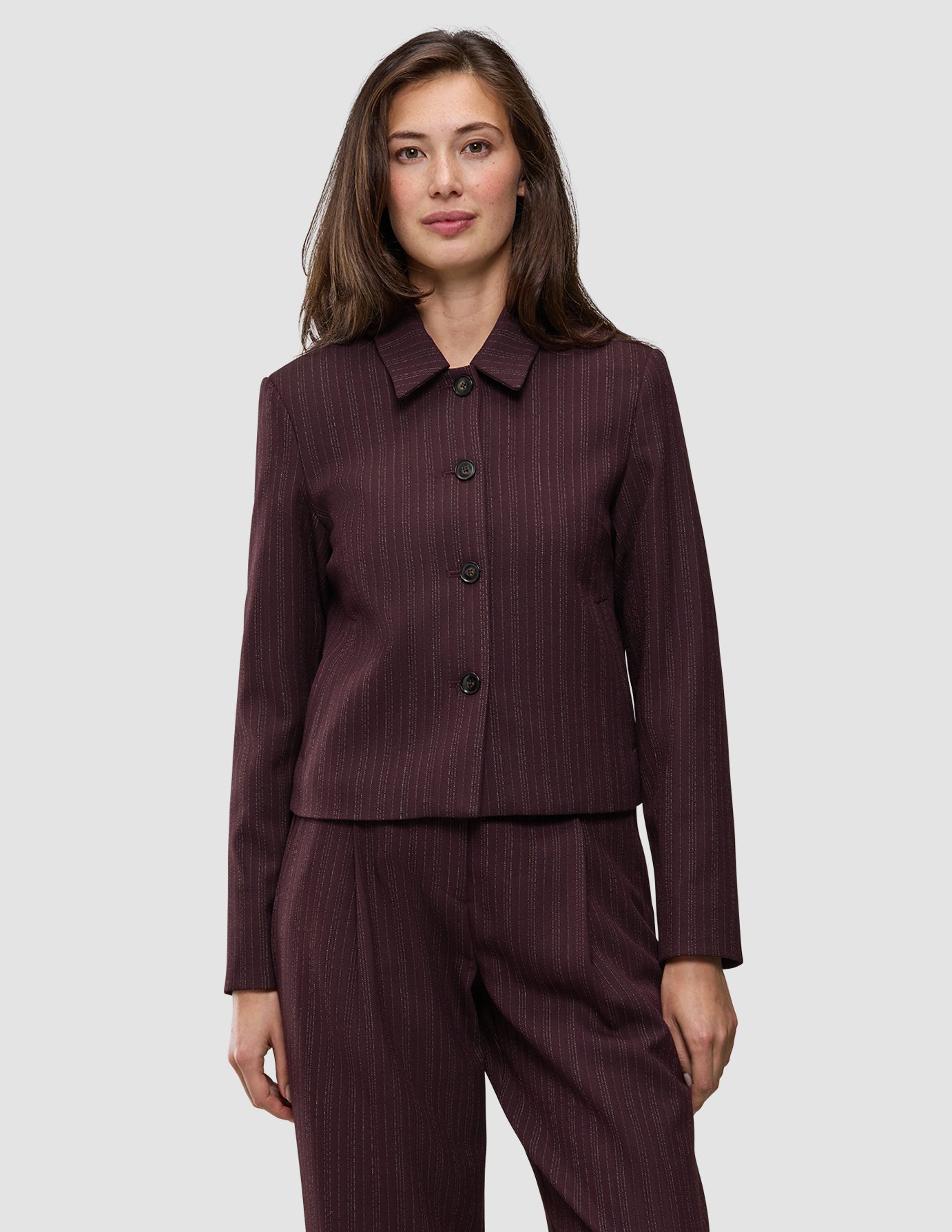 A woman wears the Women Serene Short Jacket Berry Pinstripe, a berry-colored, button-front short jacket in a Scandinavian design with thin vertical pinstripes.