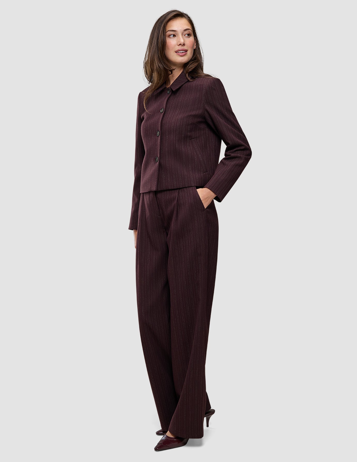 A woman wears a Women Serene Short Jacket Berry Pinstripe in a dark berry-colored Scandinavian design, paired with matching wide-leg trousers.