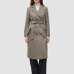 A woman wears a Women Wool Coat Greige, a double-breasted, knee-length grey wool coat with a belted waist over a white shirt and black trousers.