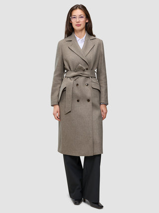 A woman wears a Women Wool Coat Greige, a double-breasted, knee-length grey wool coat with a belted waist over a white shirt and black trousers.