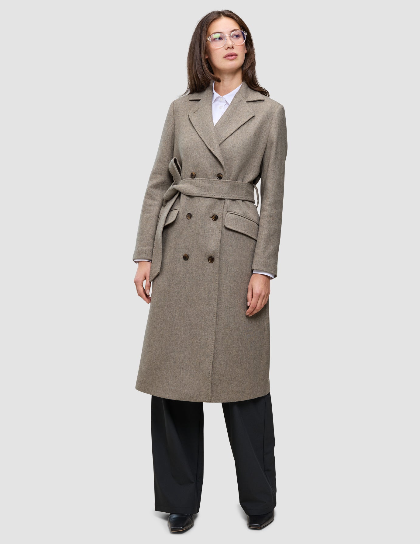 A model wears a Women Wool Coat Greige in a double-breasted Scandinavian design with a knee-length cut and matching tie belt over a white shirt and black trousers.