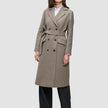 A model wears a Women Wool Coat Greige in a double-breasted Scandinavian design with a knee-length cut and matching tie belt over a white shirt and black trousers.