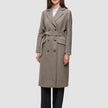 A woman wears a Women Wool Coat Greige, a long, double-breasted grey-brown wool coat with a matching tie belt over a white shirt and black trousers, showcasing minimalist Scandinavian design.