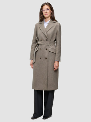 A woman wears a Women Wool Coat Greige, a long, double-breasted grey-brown wool coat with a matching tie belt over a white shirt and black trousers, showcasing minimalist Scandinavian design.