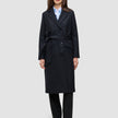A woman wears a handmade-in-Turkey Women’s Wool Coat Black in a double-breasted style with a belted waist over a light blue collared shirt and black trousers.