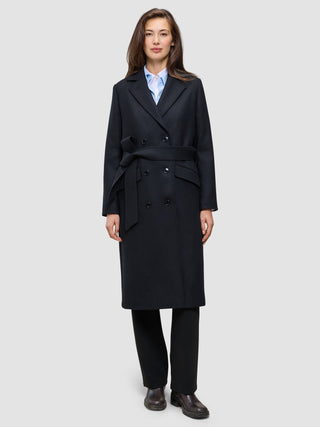 A woman wears a handmade-in-Turkey Women’s Wool Coat Black in a double-breasted style with a belted waist over a light blue collared shirt and black trousers.