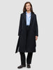 A woman wearing a Women’s Wool Coat Black in a long, double-breasted Scandinavian design over a light blue button-down shirt and black trousers.