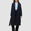 A woman wearing a Women’s Wool Coat Black in a long, double-breasted Scandinavian design over a light blue button-down shirt and black trousers.