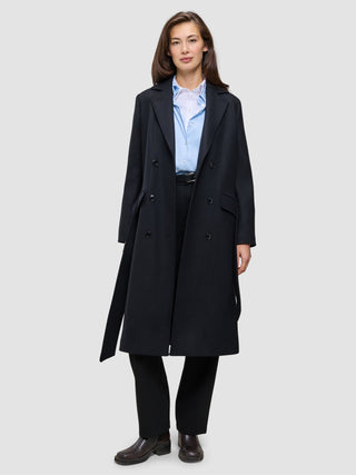 A woman wearing a Women’s Wool Coat Black in a long, double-breasted Scandinavian design over a light blue button-down shirt and black trousers.