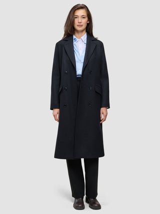 A person wearing a Women’s Wool Coat Black in a double-breasted Scandinavian design over a light blue collared shirt and black trousers.