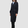 A woman wearing a Women’s Wool Coat Black in a Scandinavian-design double-breasted style with a tie waist over a light blue shirt and black trousers.