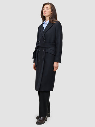 A woman wearing a Women’s Wool Coat Black in a Scandinavian-design double-breasted style with a tie waist over a light blue shirt and black trousers.
