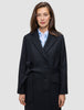 A woman wearing a Women’s Wool Coat Black in a Scandinavian-design double-breasted style with a tie waist over a light blue collared shirt.