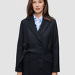 A woman wearing a Women’s Wool Coat Black in a Scandinavian-design double-breasted style with a tie waist over a light blue collared shirt.