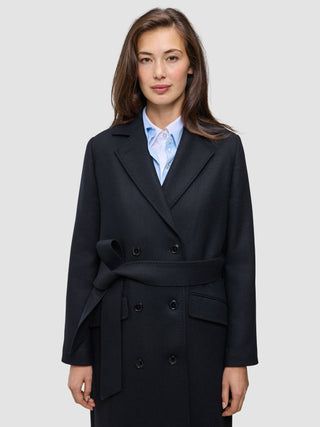 A woman wearing a Women’s Wool Coat Black in a Scandinavian-design double-breasted style with a tie waist over a light blue collared shirt.