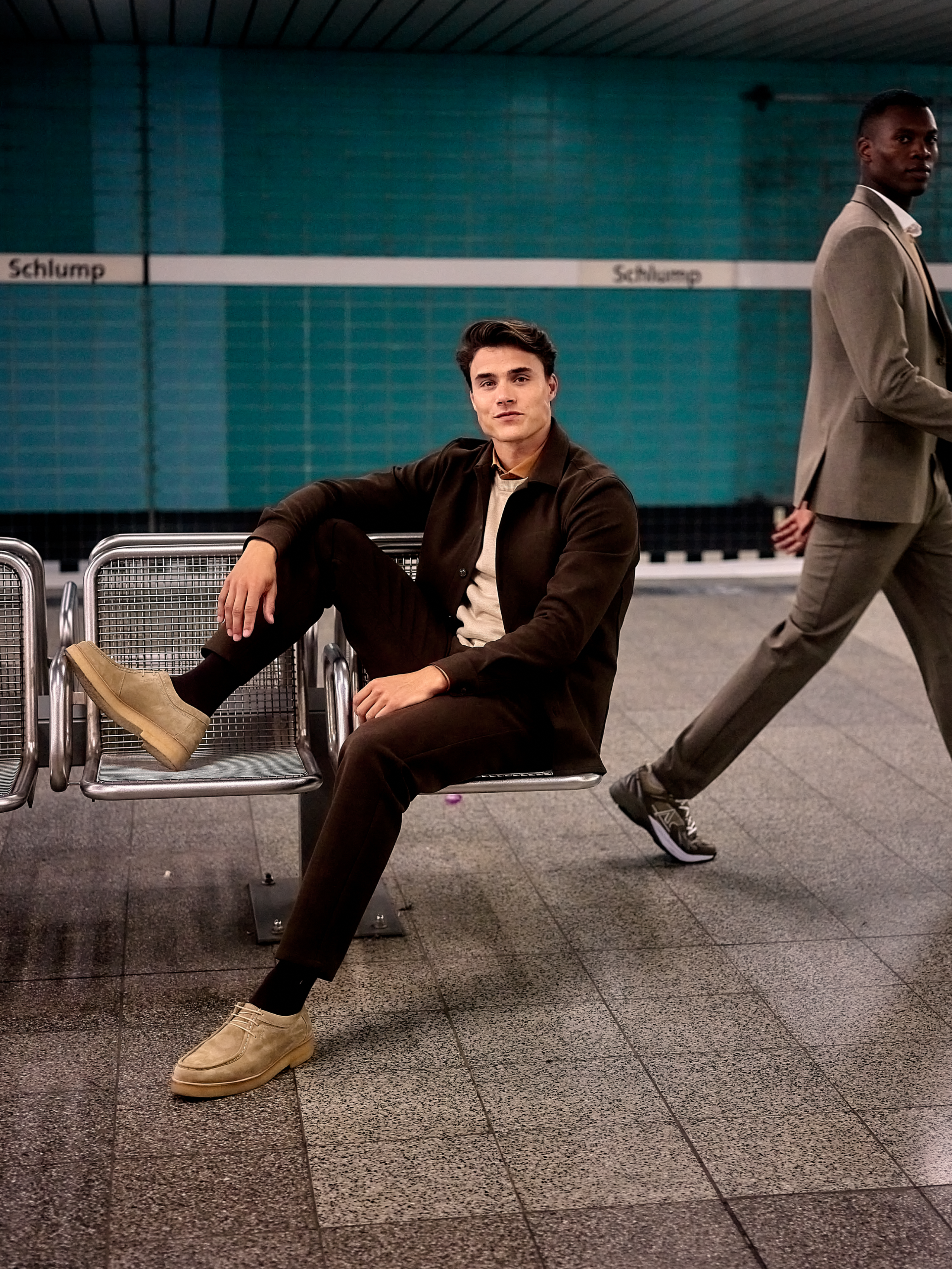 Male model wearing brown pants, beige pullover and brown overshirt sitting in an underground station