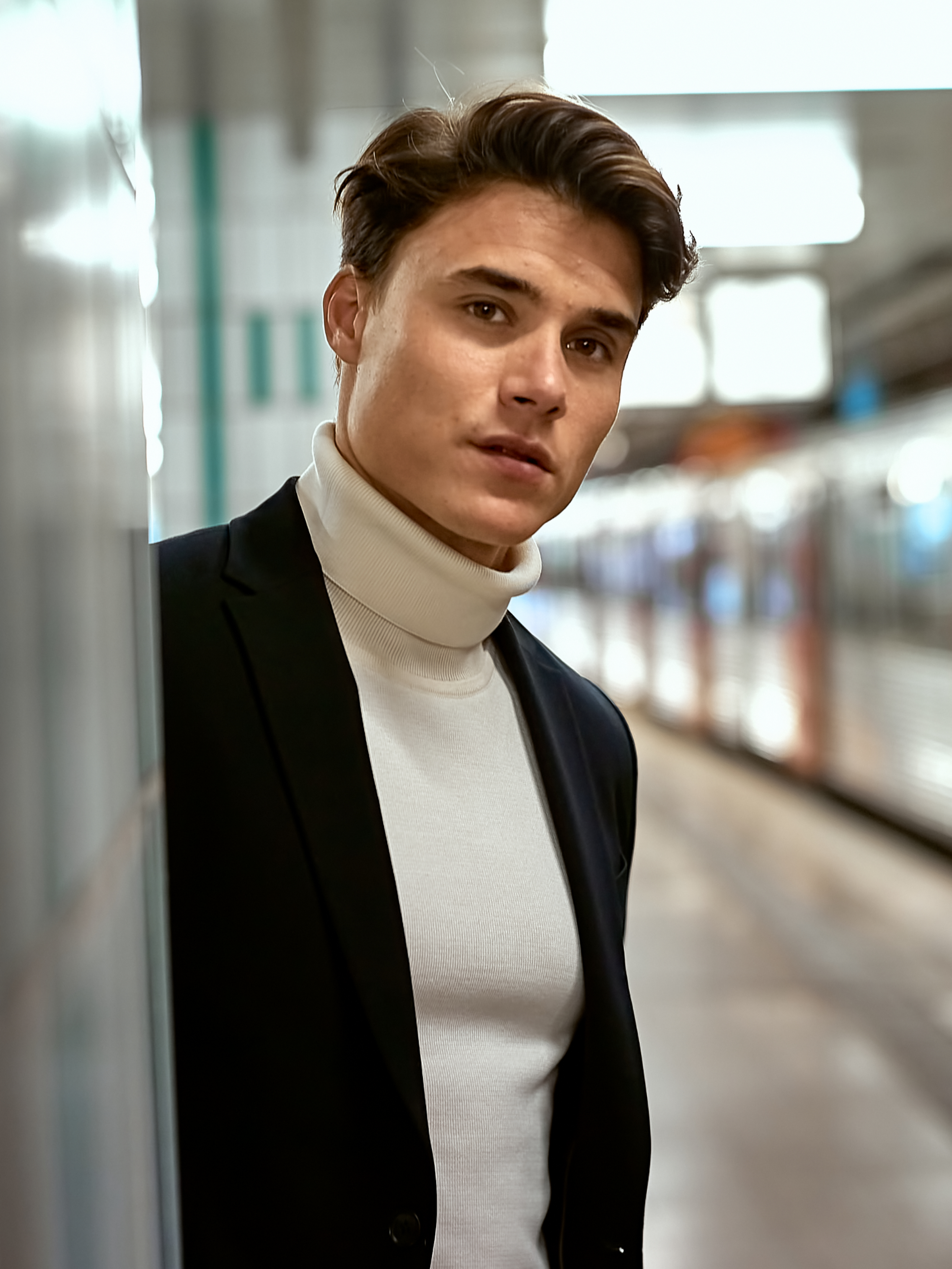 Male model wearing beige turtleneck and black blazer standing in a metro station