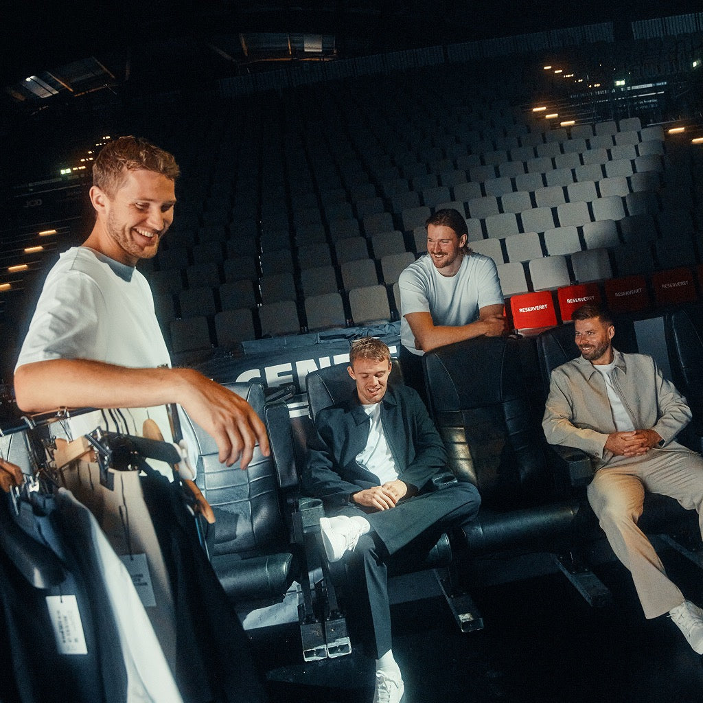 Four men interacting in a darkened theater with stadium-style seating, including one man in the foreground holding a clothing rack.