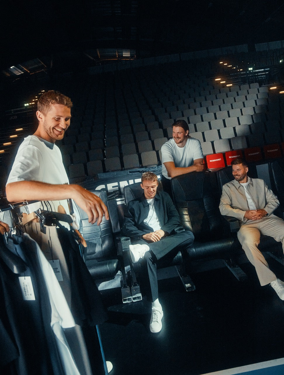 Four men in casual-smart attire gathered in a stadium seating area, with one man standing in the foreground near a clothing rack.