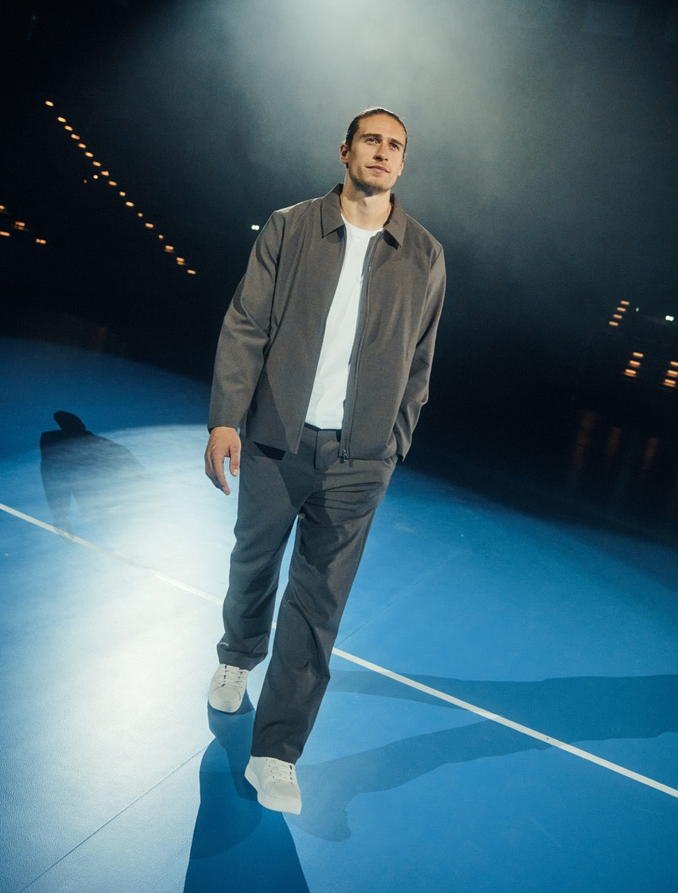 A man walking across a blue sports court wearing a grey jacket, white t-shirt, and grey trousers.