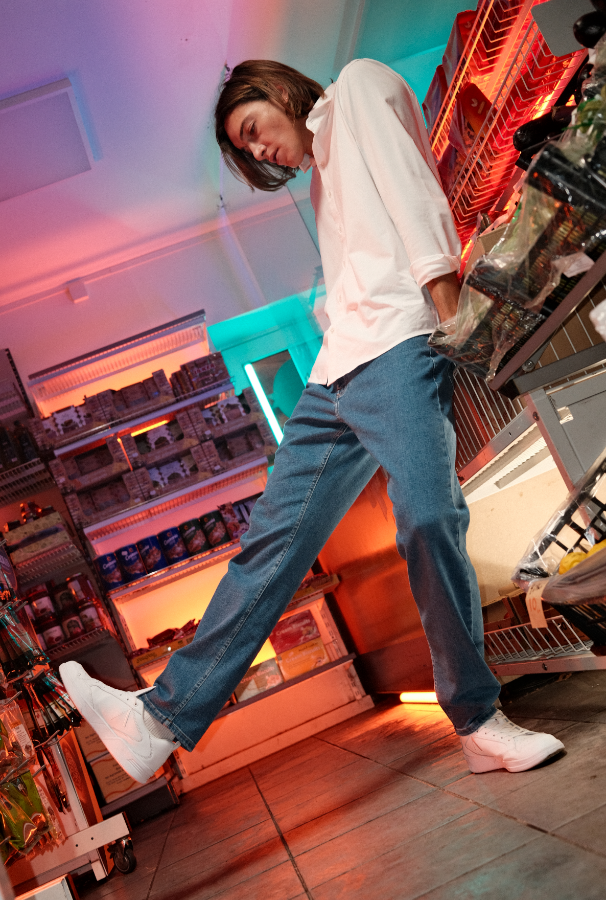 Male model wearing light blue jeans, white shirt, white sneakers lifting one leg up showing stretch of jeans