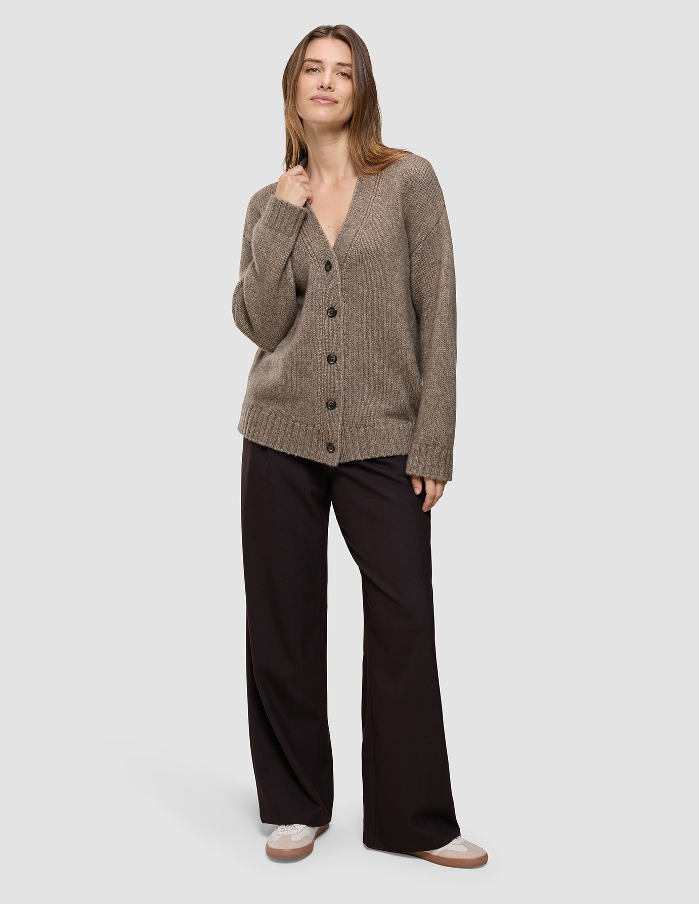 A woman wearing a Women’s Chunky Soft Wool Knit Cardigan Taupe Melange, a cozy V-neck button-down knit cardigan paired with dark wide-leg trousers.