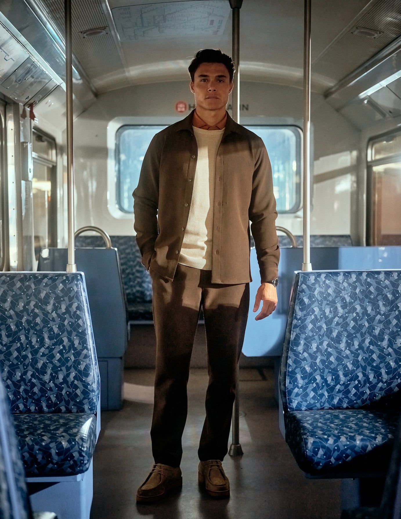 Male model wearing brown pants, beige pullover and brown overshirt standing in a bus