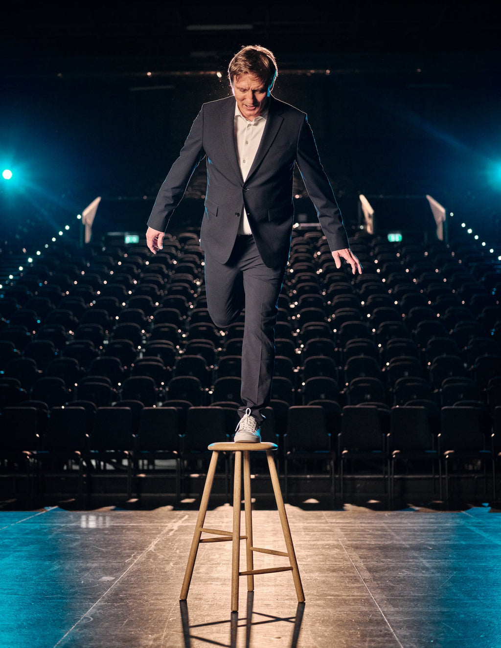 Oliver Masucci wearing a grey suit, white shirt and white sneakers balancing one foot on a chair in a theater